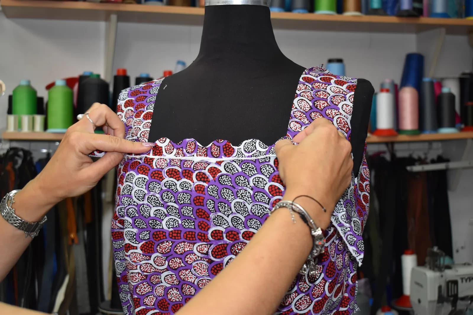 Enisa, a MadeApt tailor, pinning the neckline of a purple printed dress on a dress form in her Sarajevo studio, shelves of thread behind her