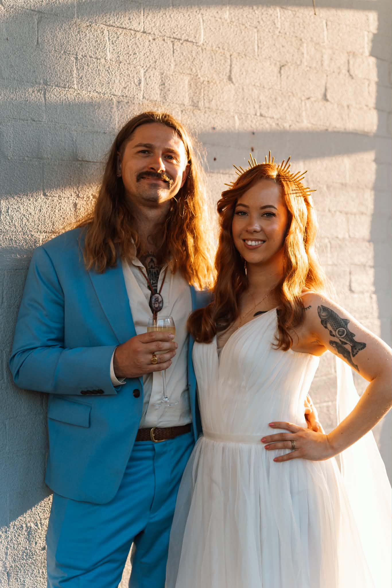 A bride in a white tulle V-neck gown and gold crown standing with her partner in a blue suit against a sunlit brick wall