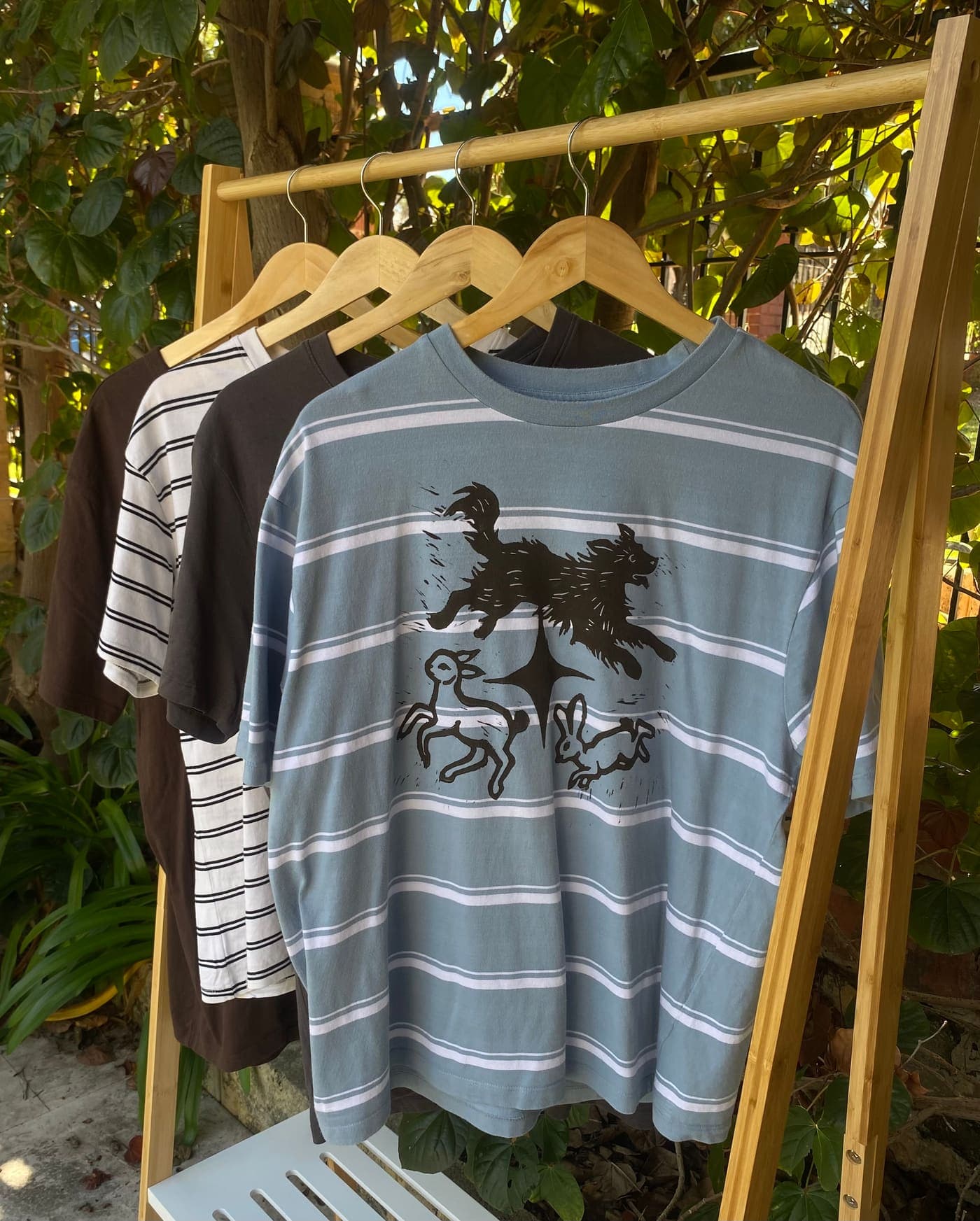 A light blue striped tee hand-printed with a running dog, lamb and hare on a wooden rack in the garden
