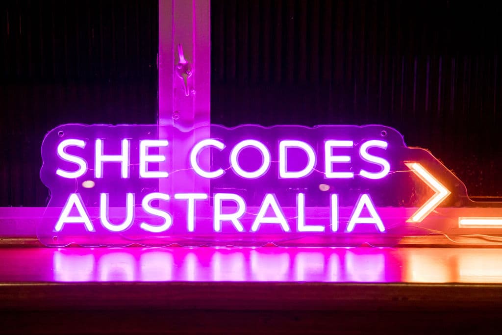 A neon sign reading She Codes Australia, glowing violet