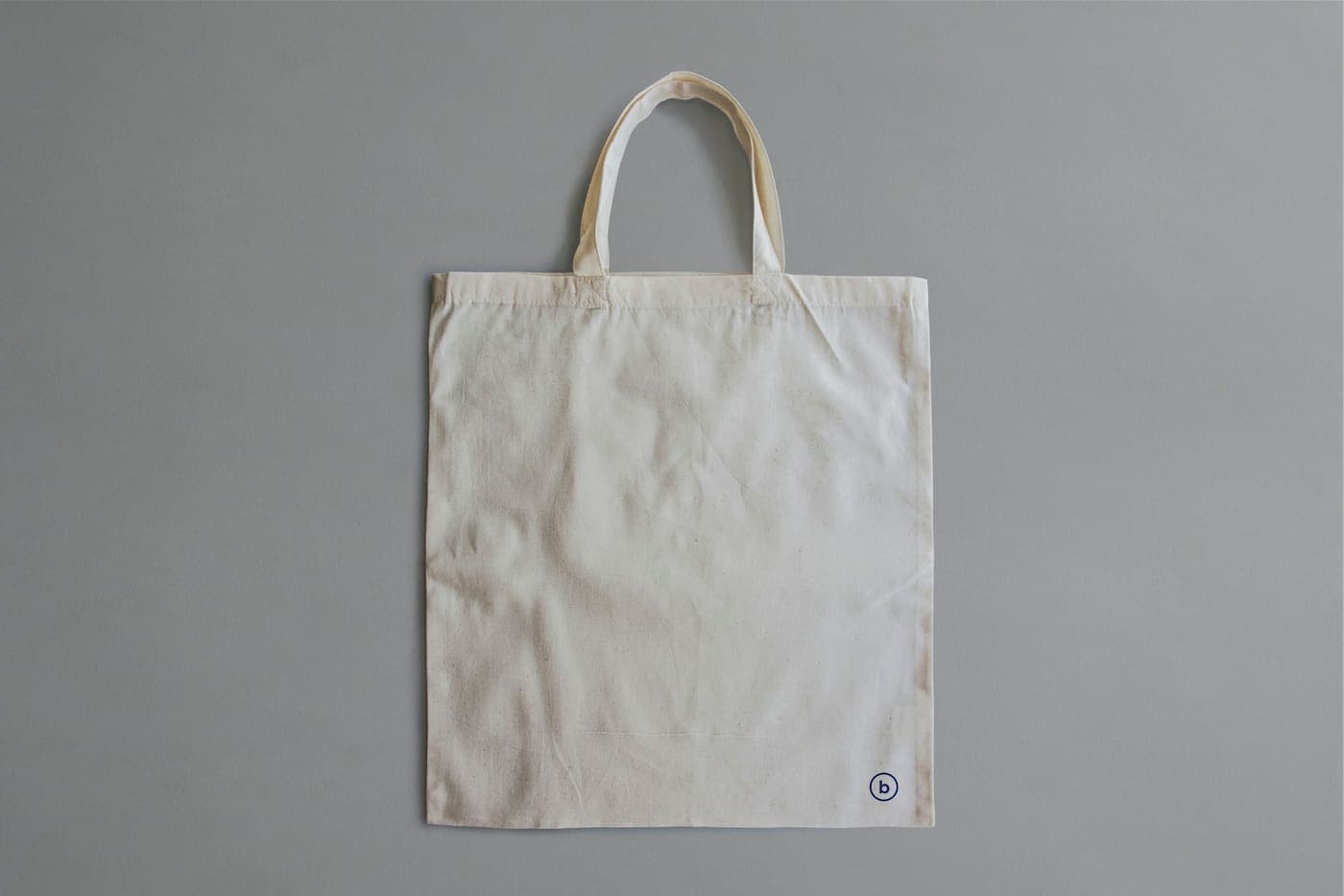 A plain natural canvas tote bag laid flat on a grey surface, unprinted and ready for artwork or embroidery