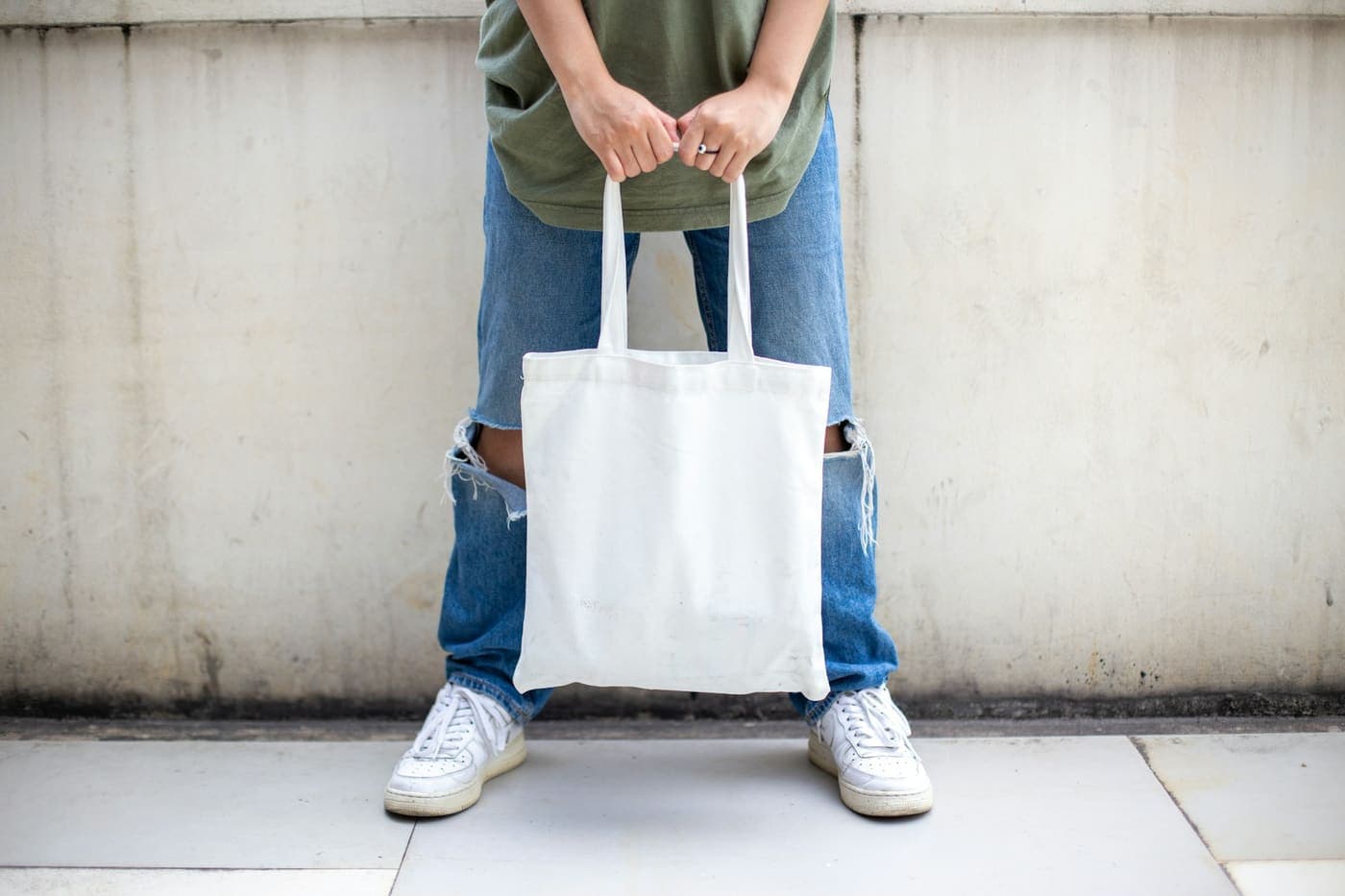 A person in blue jeans and white sneakers holding a plain white canvas tote bag by its handles